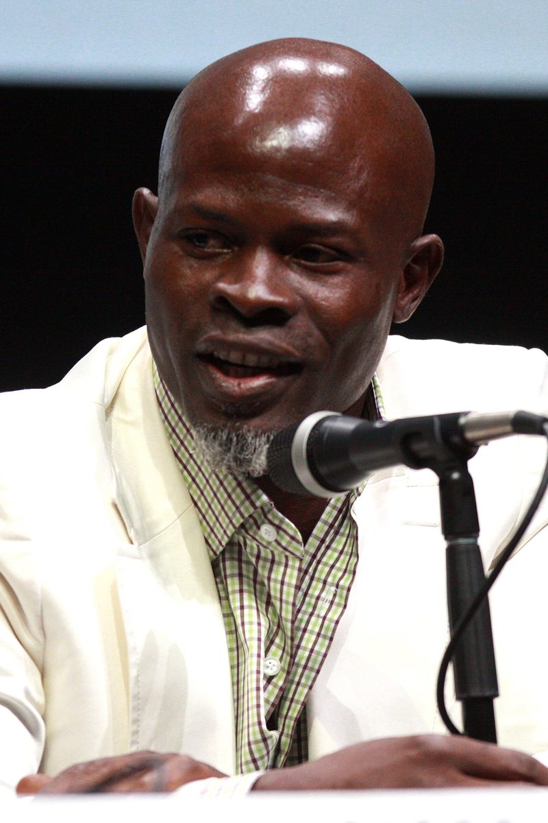 Djimon Hounsou in a cream jacket speaking into a microphone at a panel