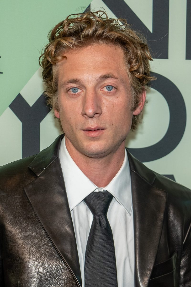 Jeremy Allen White with curly hair in a black leather jacket, white shirt and black tie