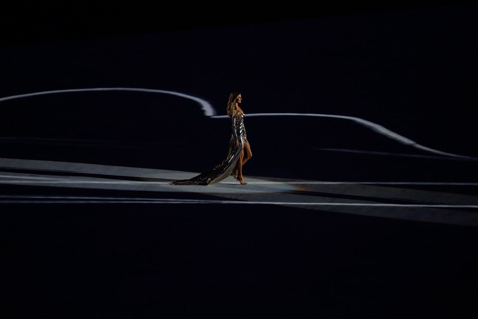 Gisele Bündchen in a long silver gown walking alone across the dark stadium floor at the Rio 2016 Olympic opening ceremony