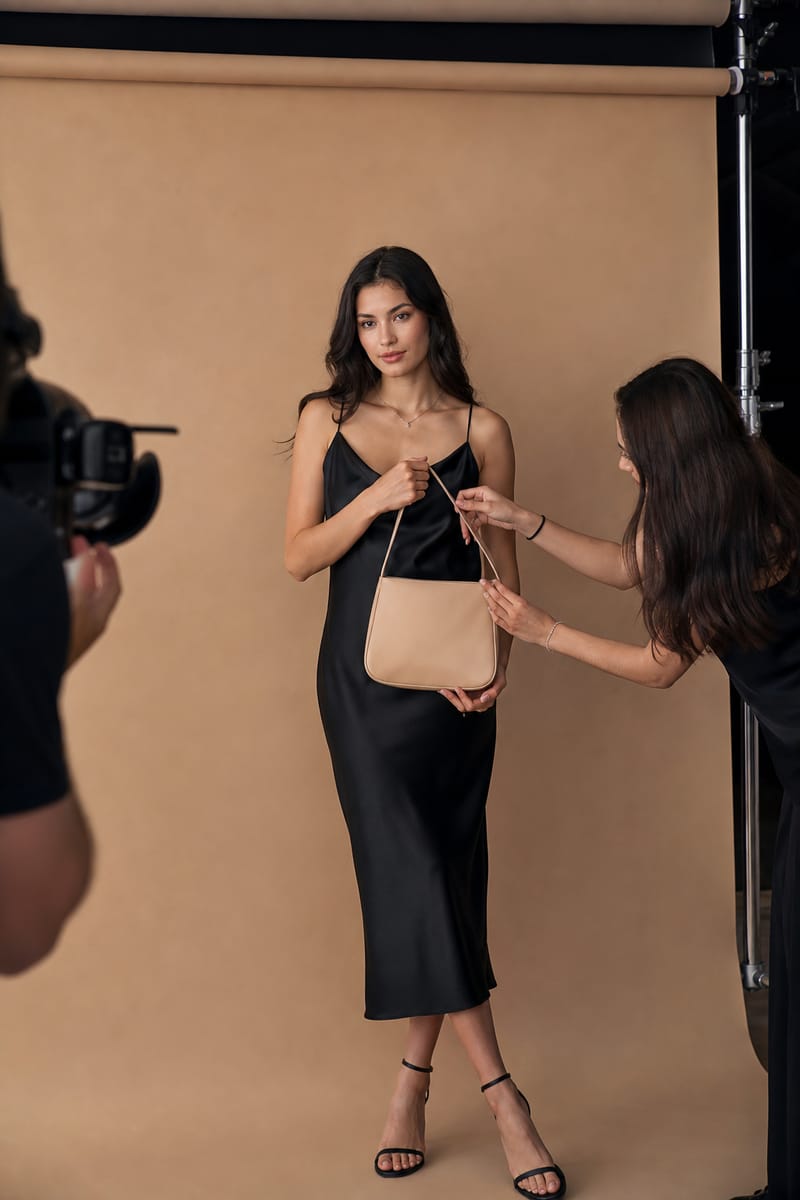Creator in a black dress poses with a leather handbag on a brand shoot while a camera operator and a stylist work at the edges of the frame