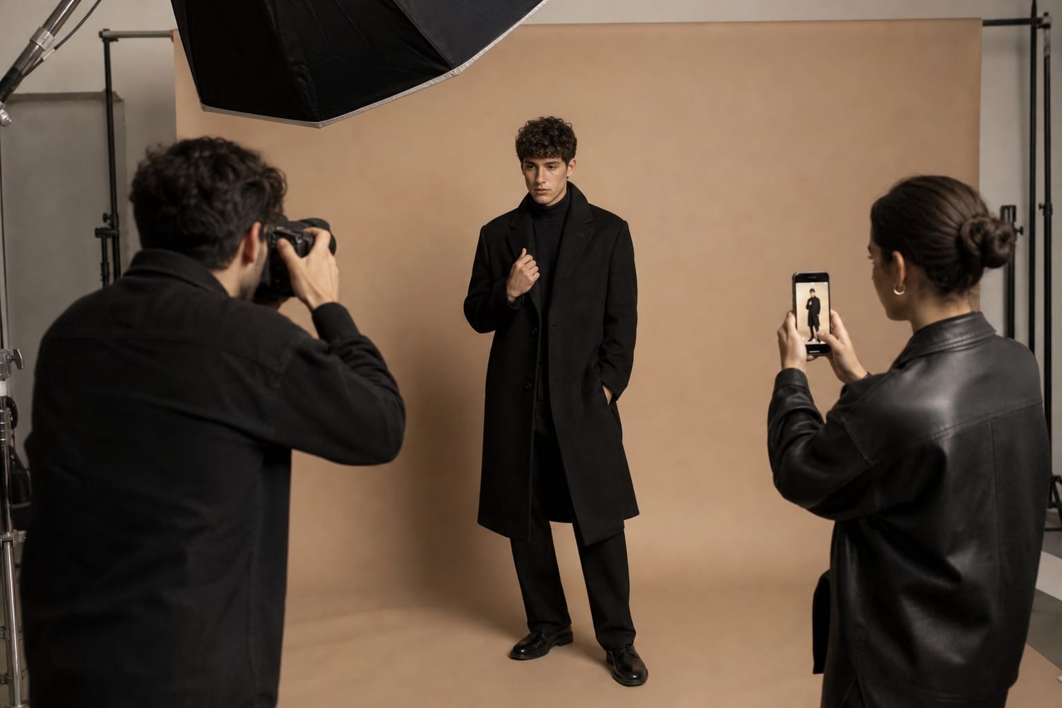 Male model in a black coat poses for a photographer in a studio while an assistant films behind-the-scenes footage on a phone