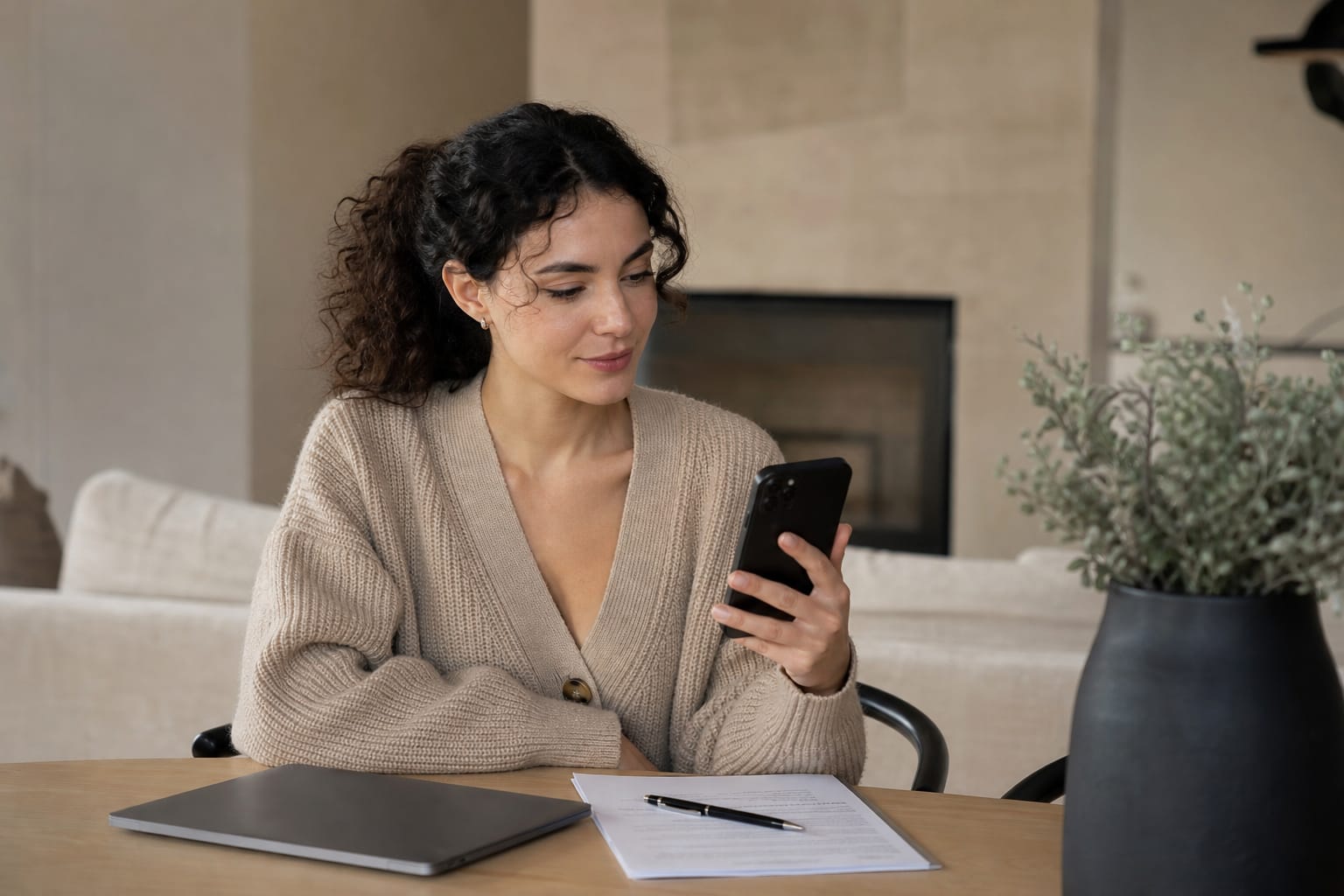Woman at a table reviewing her video on a phone, with a laptop and a printed contract beside her