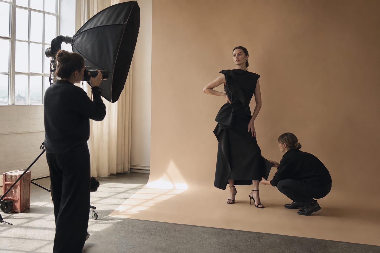 A magazine fashion shoot in a studio: a model in a sculptural black dress poses while a photographer and a stylist work beside her