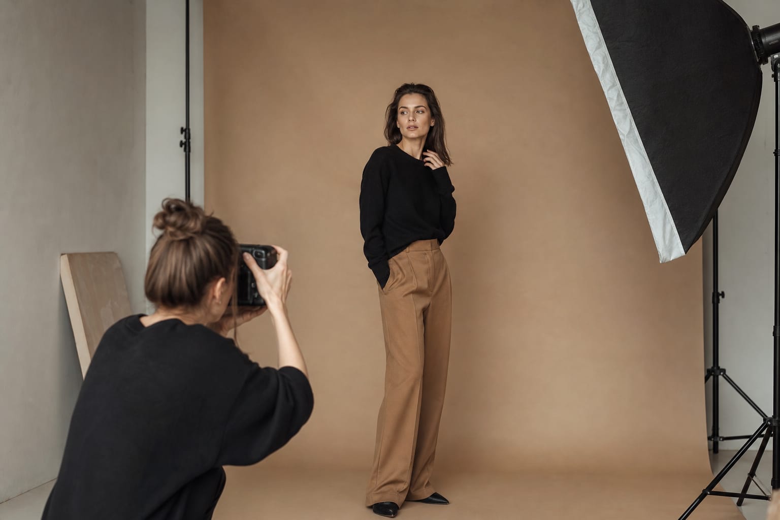 A print model posing for a photographer in a studio with a light stand and a warm beige backdrop