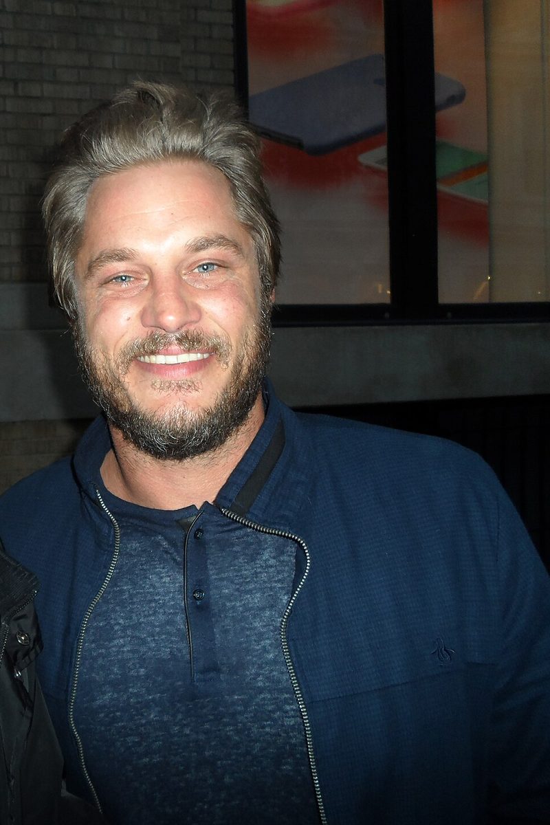 Travis Fimmel smiling outdoors at night in 2016, with longer swept-back hair and a short beard, wearing a navy zip jacket