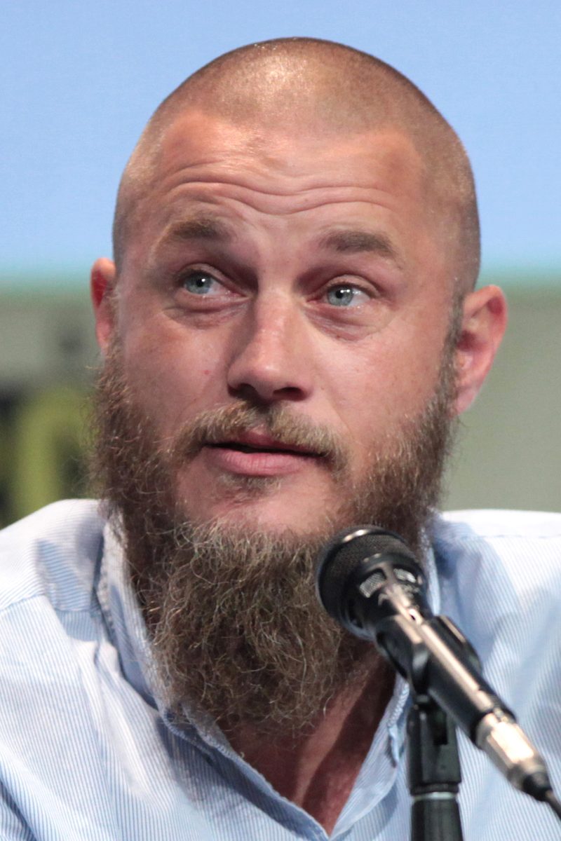 Travis Fimmel with a shaved head and a long full beard speaking at a microphone at San Diego Comic-Con in 2015