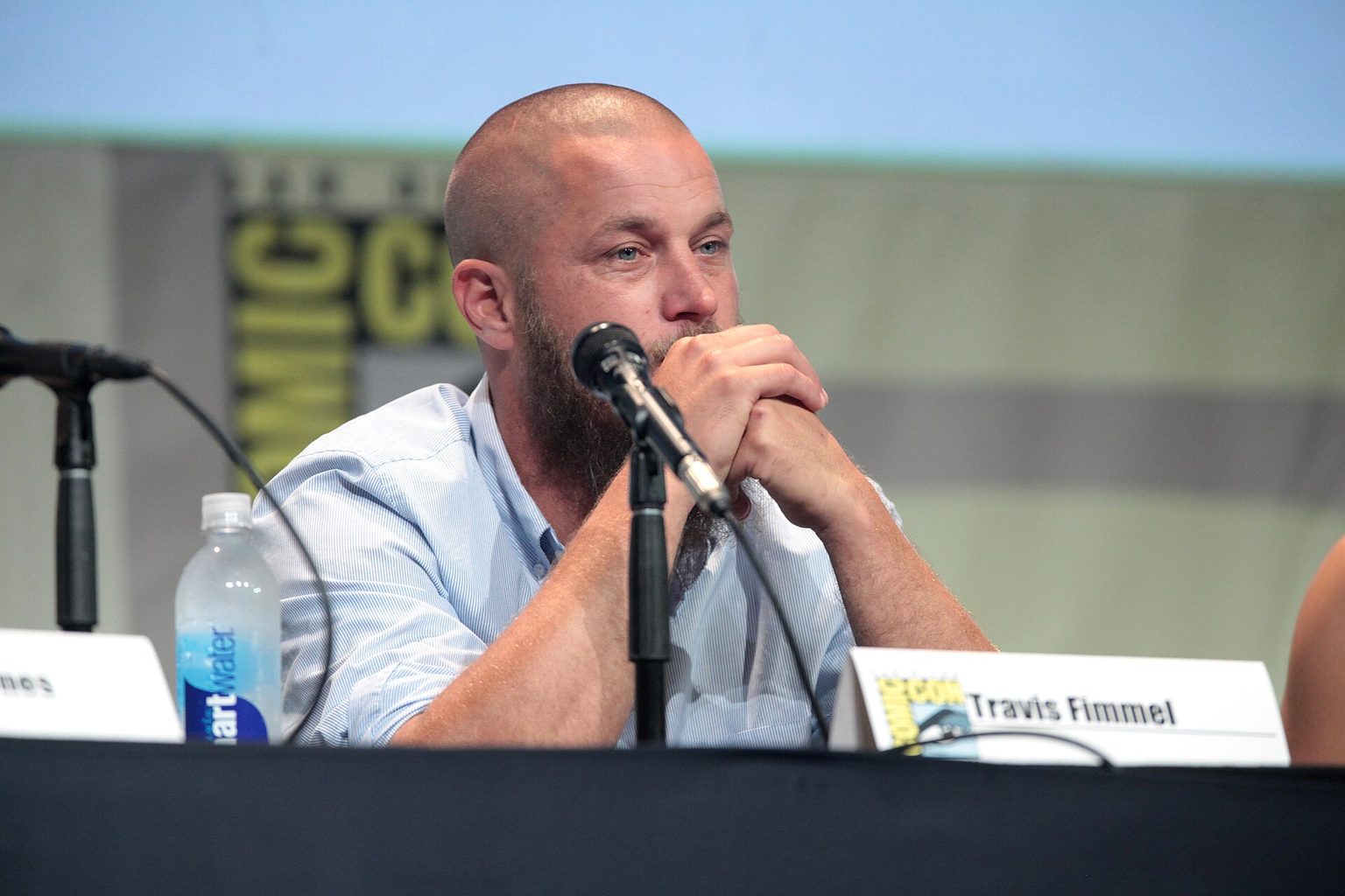 Travis Fimmel with a long beard and light blue shirt speaking at a panel table with a microphone